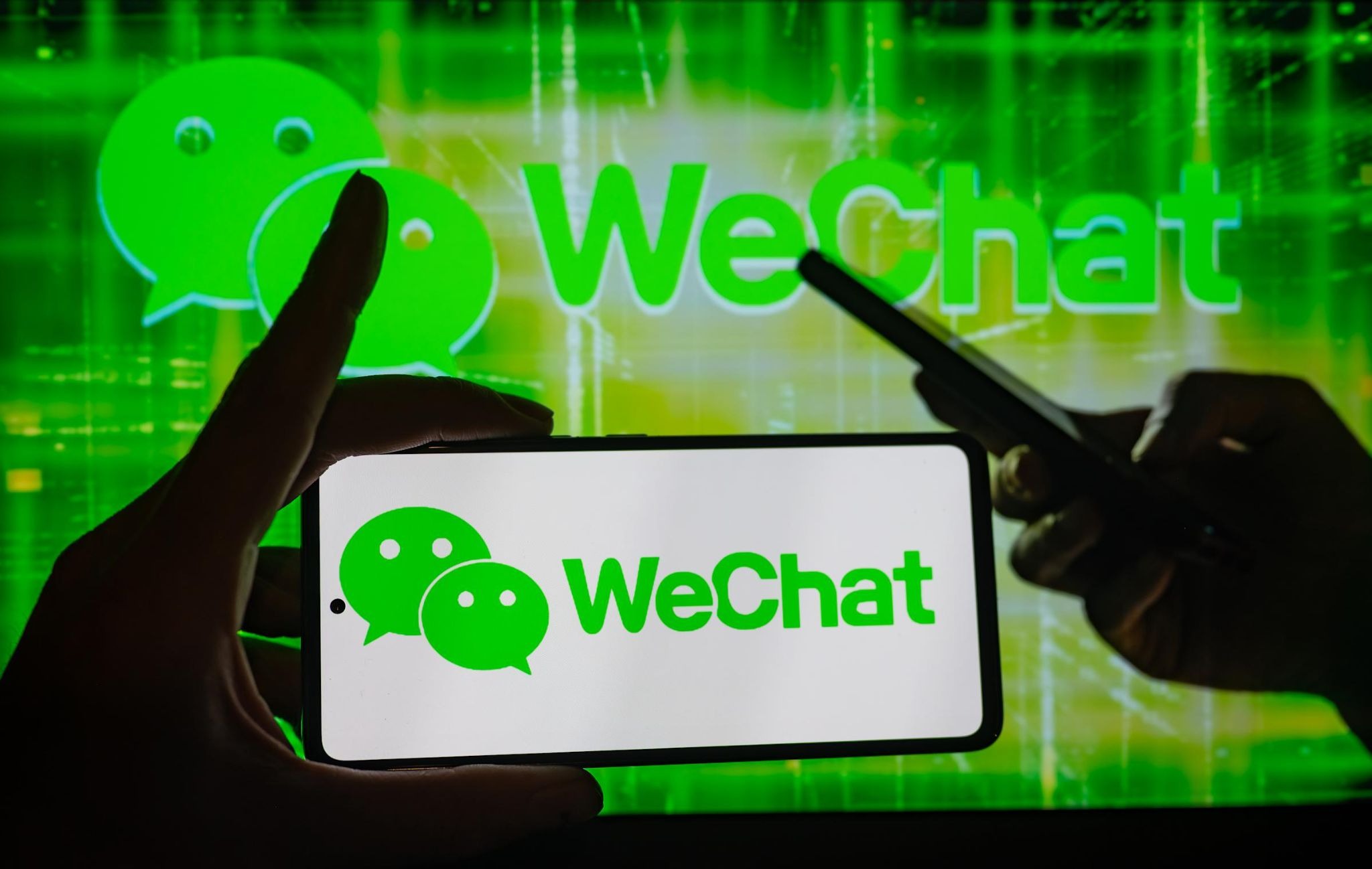 A hand holds a smartphone displaying the green WeChat logo and text, set against a blurry, glowing green background that features the same logo and another person holding a phone.